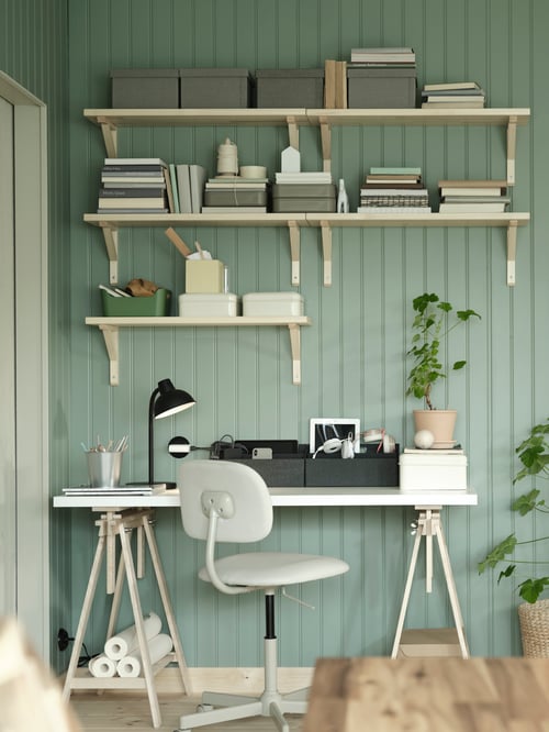 A workspace featuring KVARNVIK storage boxes with lids placed on white shelves, and a swivel chair placed next to a trestle.