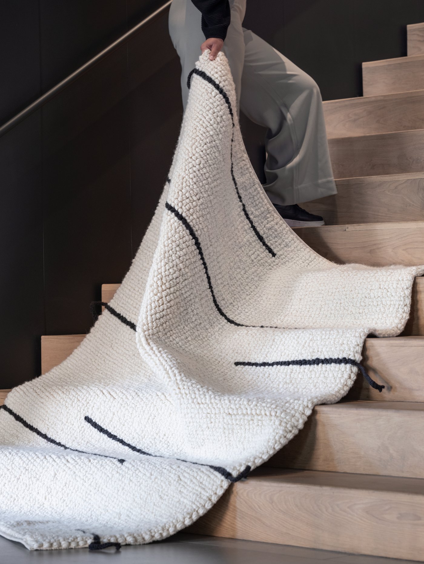 A person standing in a staircase holds a corner of a handwoven white and black LOKALT wool rug.