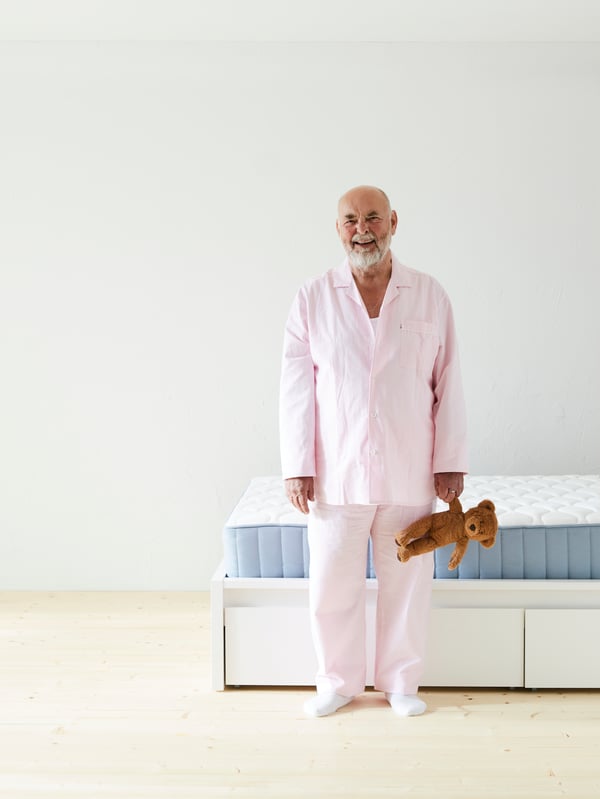 A man in pajamas stands next to a bed and looks happily at the camera