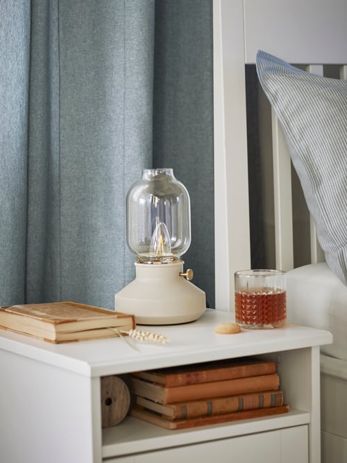 Bedroom setting with a focused shot of a table lamp, book, and glass with liquid on a nightstand containing multiple books.