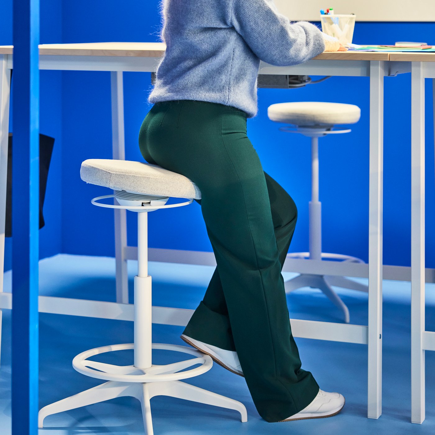 Person sittande på en hög pall vid ett högt skrivbord i en blåtonad arbetsyta, med en annan pall och ett bord synliga i närheten.
