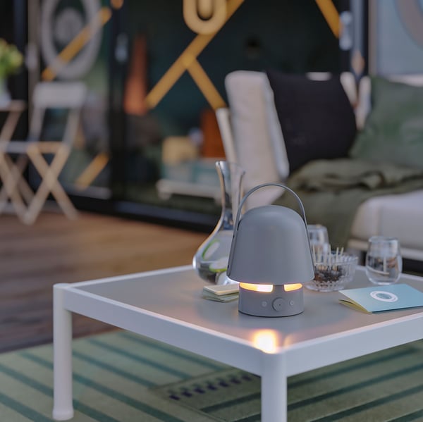 Solen går ner över ett utomhusområde. En tänd VAPPEBY Bluetooth-högtalarlampa står på ett SEGERÖN soffbord.
