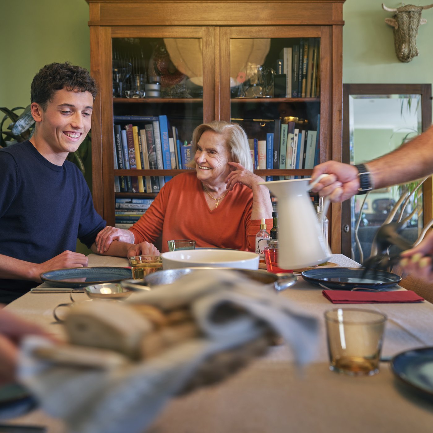 A young man and an older woman sits by a dining table while people around them serves dinner.