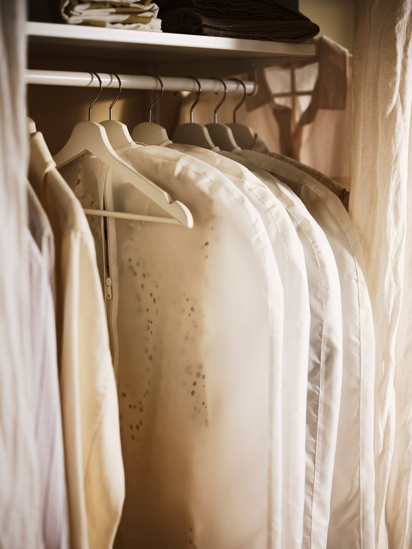 Varias perchas blancas con fundas para ropa blancas cuelgan de una barra para ropa que hay debajo de un estante blanco.