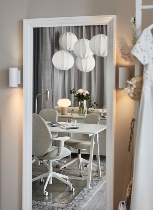 A mirror that reflects a dining room containing multiple, white pendant lamps, office chairs, a lit lamp, and a desk.