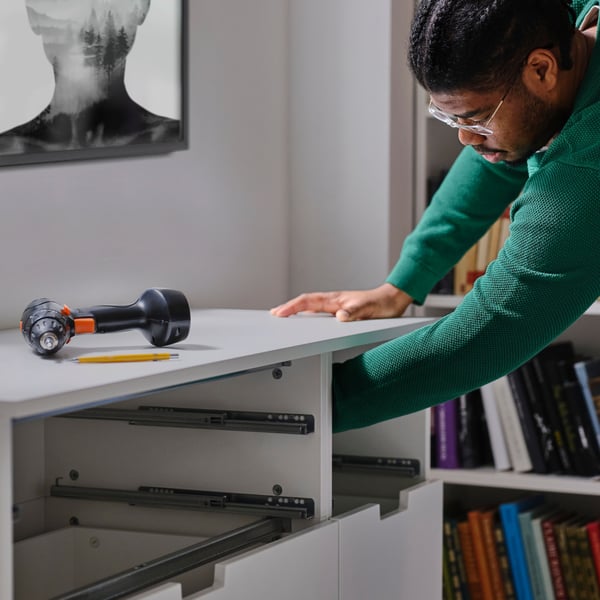 A man in green sweater, assembling a chest of drawers, making sure to anchor it to the wall to avoid tip-over accidents.