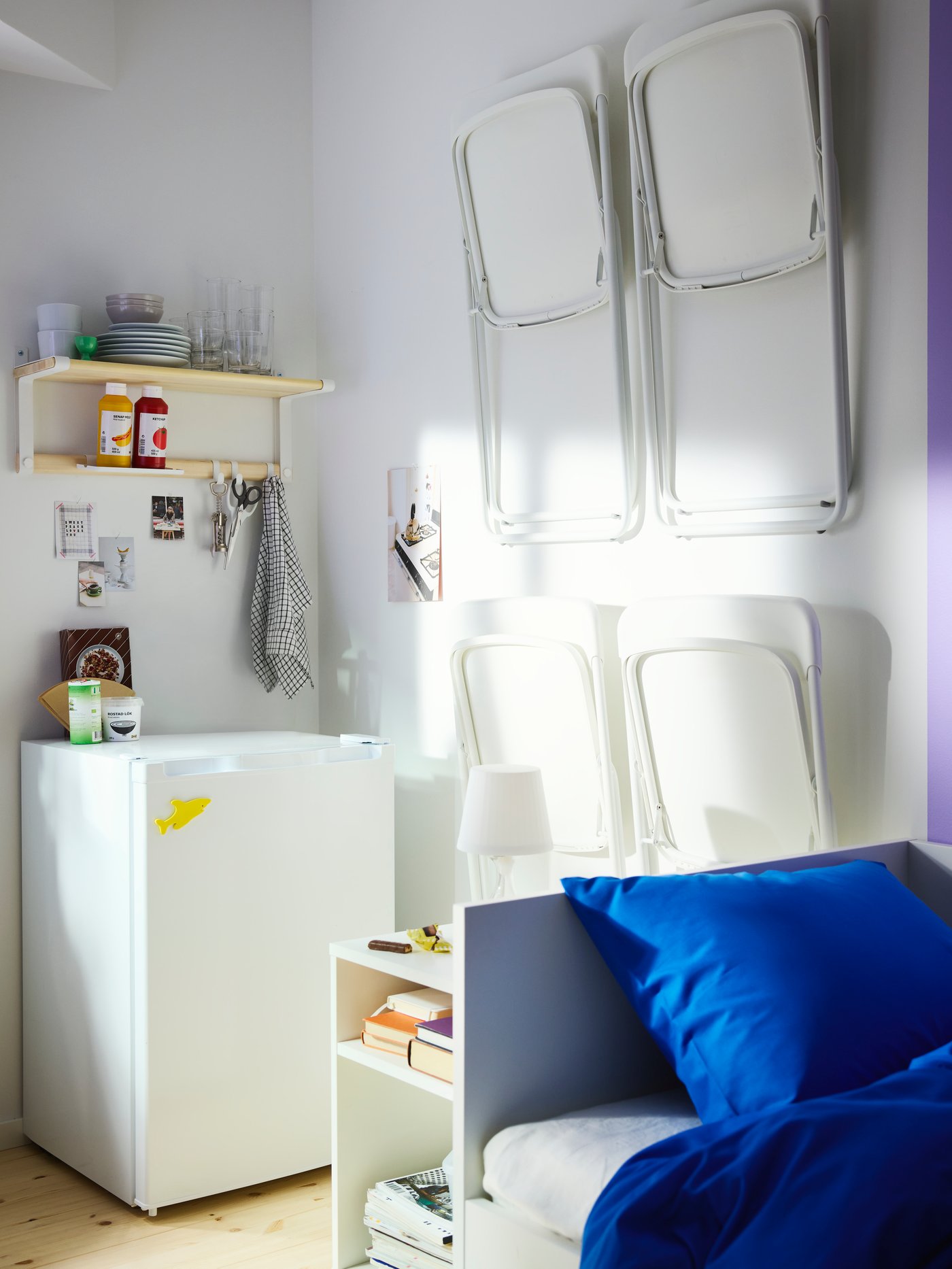 Čtyři bílé skládací židle VIHALS visí na stěně mezi postelí a malou ledničkou s policí nad ní.