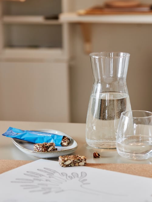 Workspace with a glass carafe with water, near a glass, a drawing, a snack on a plate, placed on a table, and furniture.