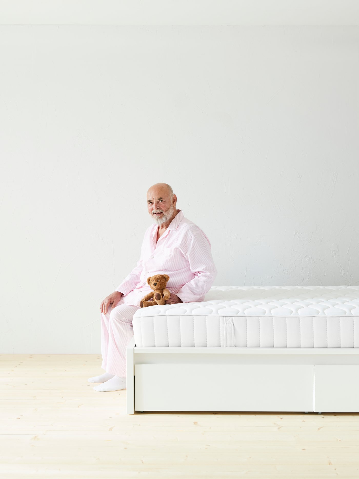 Eine Person in rosa Schlafkleidung sitzt auf dem Rand einer weißen Matratze in einem hellen Raum und hält ein kleines Stofftier auf dem Schoß.