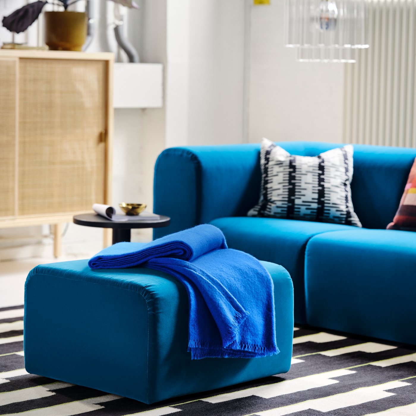 Modernt vardagsrum med blå STOCKHOLM soffa och sittpuff, mönstrade kuddar, blå filt, sidobord med guldfärgad skål och svartvit randig matta.