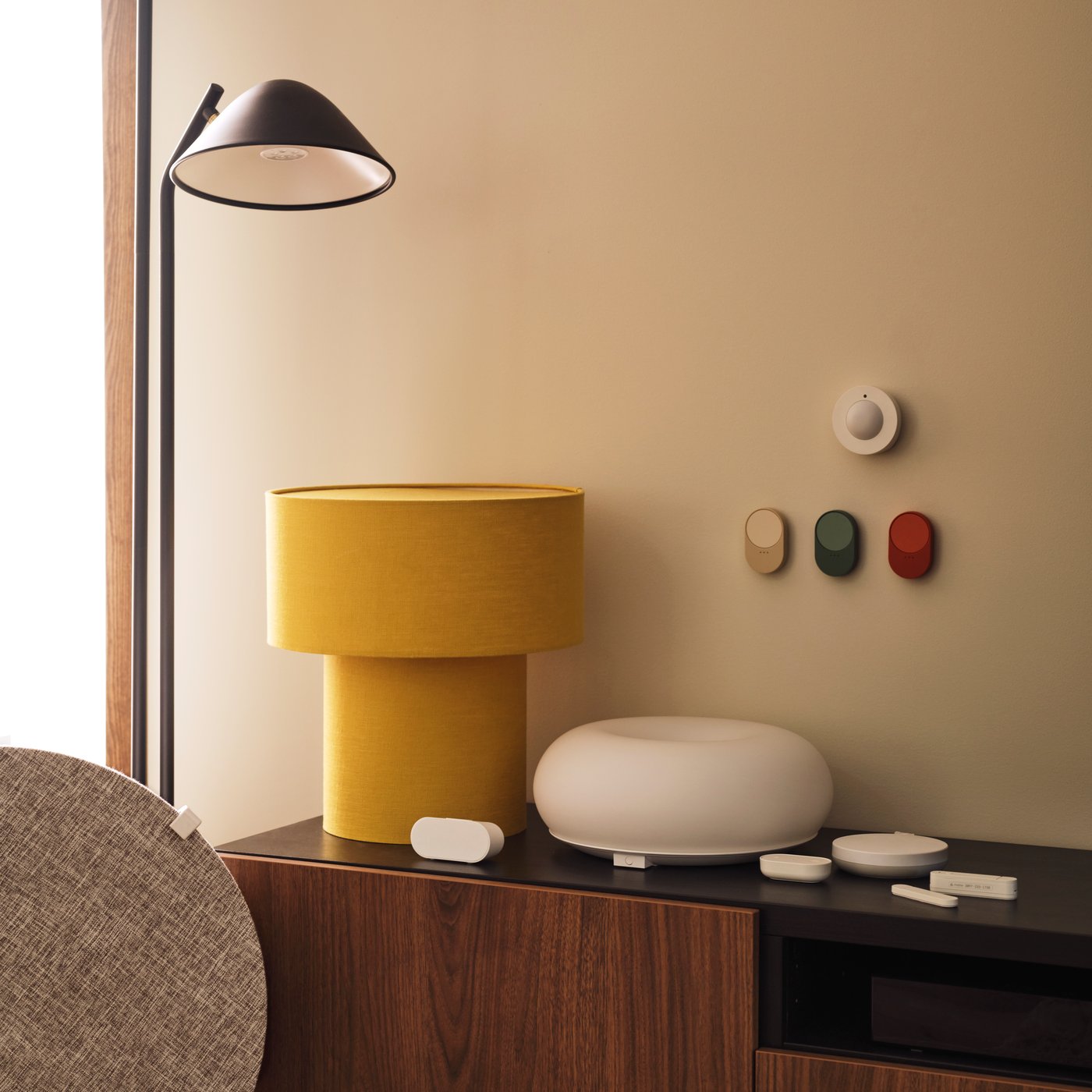 A MYGGSPRAY wireless motion sensor and three BILRESA remote controls are mounted on a wall above a TV bench next to a lamp.