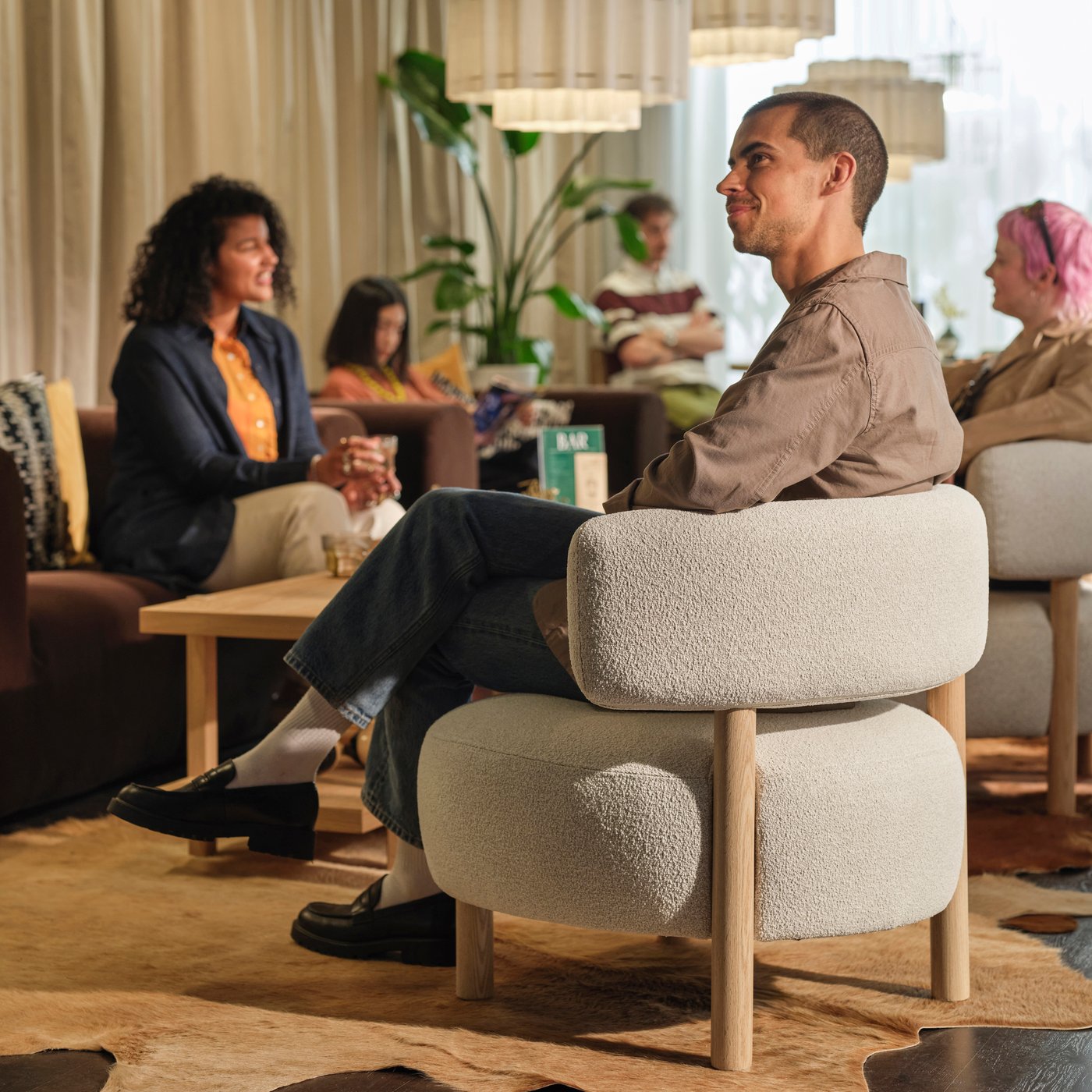 Person sittande i en stoppad fåtölj i en mysig lounge, med andra som pratar i närheten under pendellampor och bredvid ett litet bord.