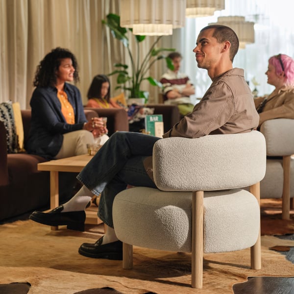 Person sittande i en stoppad fåtölj i en mysig lounge, med andra som pratar i närheten under pendellampor och bredvid ett litet bord.