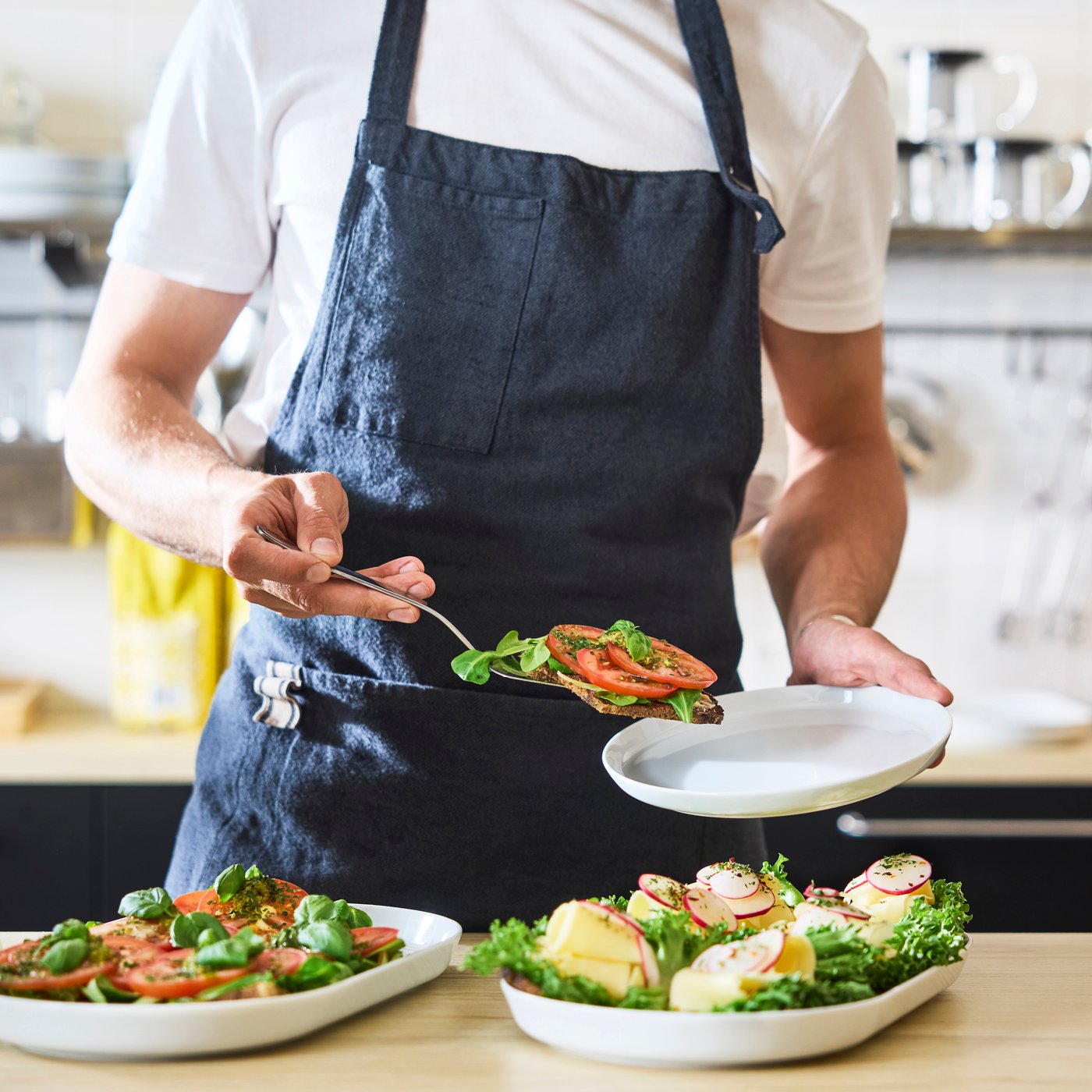 Barista lägger upp öppna smörgåsar toppade med grönsaker och tomat på en tallrik; sallader och färdigrätter arrangeras på disken.