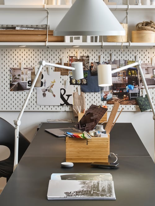 A white NYMÅNE floor and reading lamp stands by either side of a black LINNMON/ODDVALD table filled with study accessories.