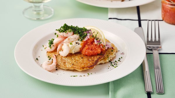 Two golden-brown rösti have shrimp, caviar and a lemon slice and they are placed on a white plate on a green tablecloth.