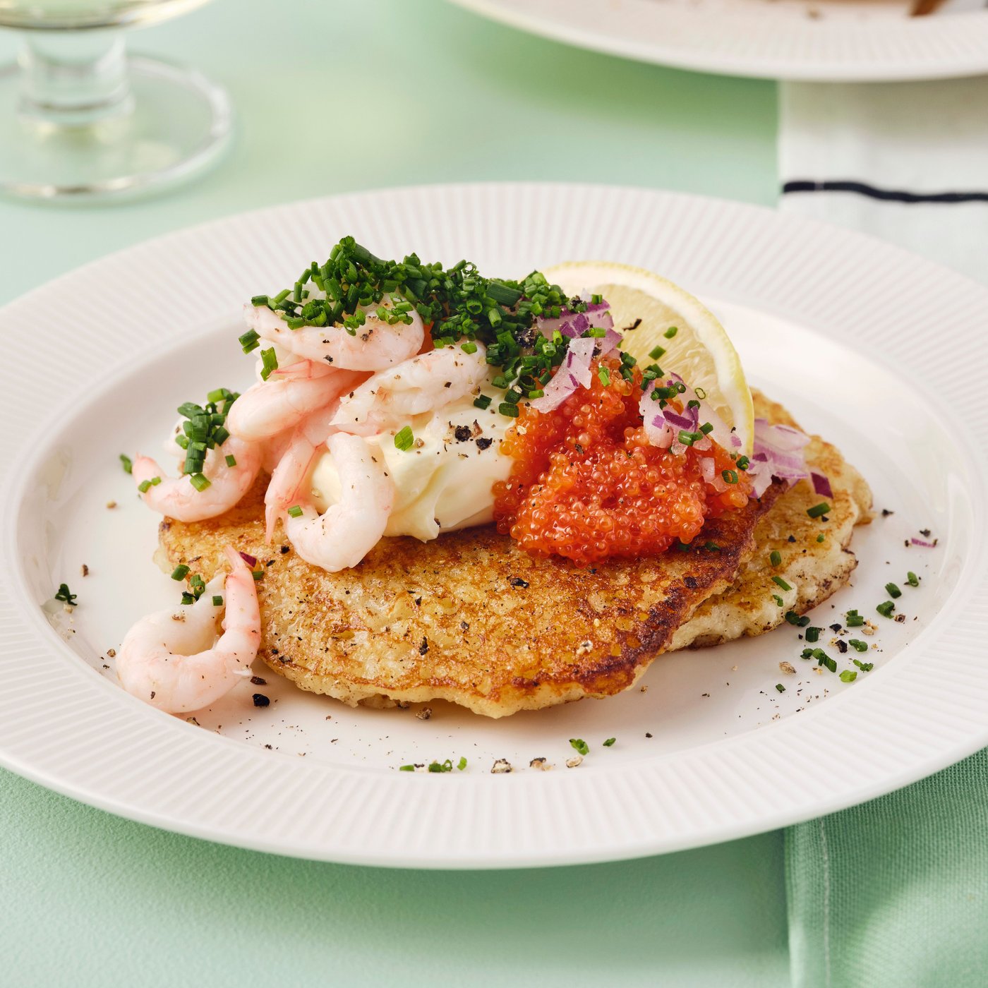 Due rösti dorati con gamberetti, caviale e una fetta di limone in un piatto bianco su una tovaglia verde.