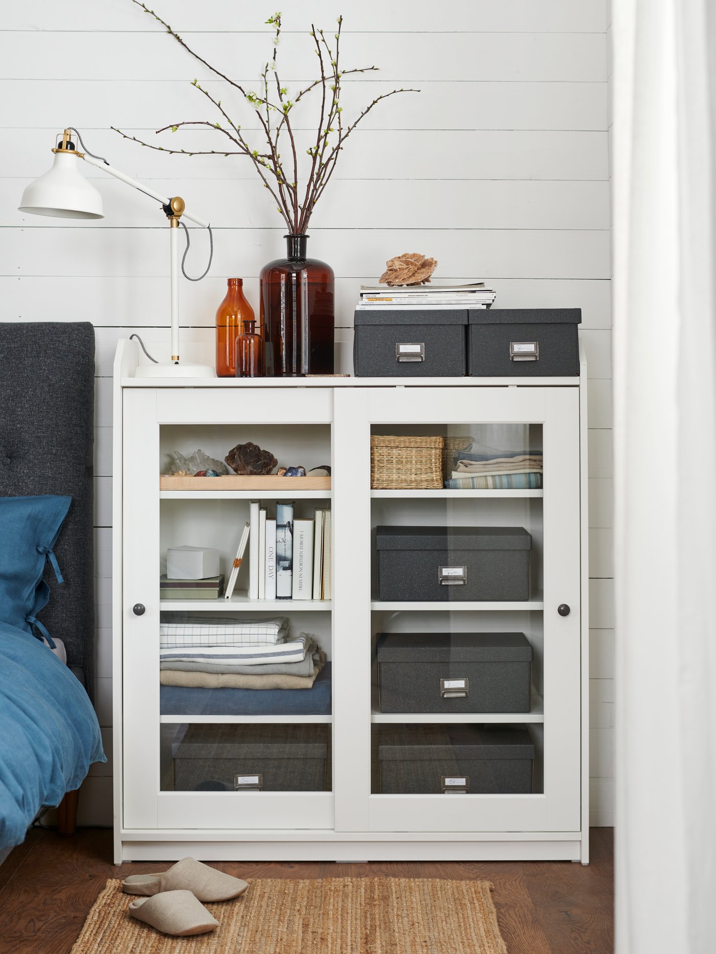 Dormitorio con vitrina blanca utilizada como mesilla de noche, con una lámpara de trabajo, cajas de almacenaje y adornos.