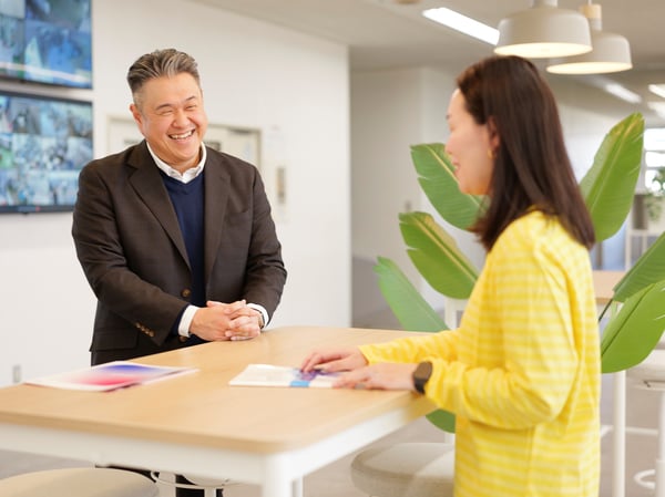 スーツ姿の人物と黄色いトップスの人物が、観葉植物のある明るいオフィスで白いテーブル越しに向かい合っている。