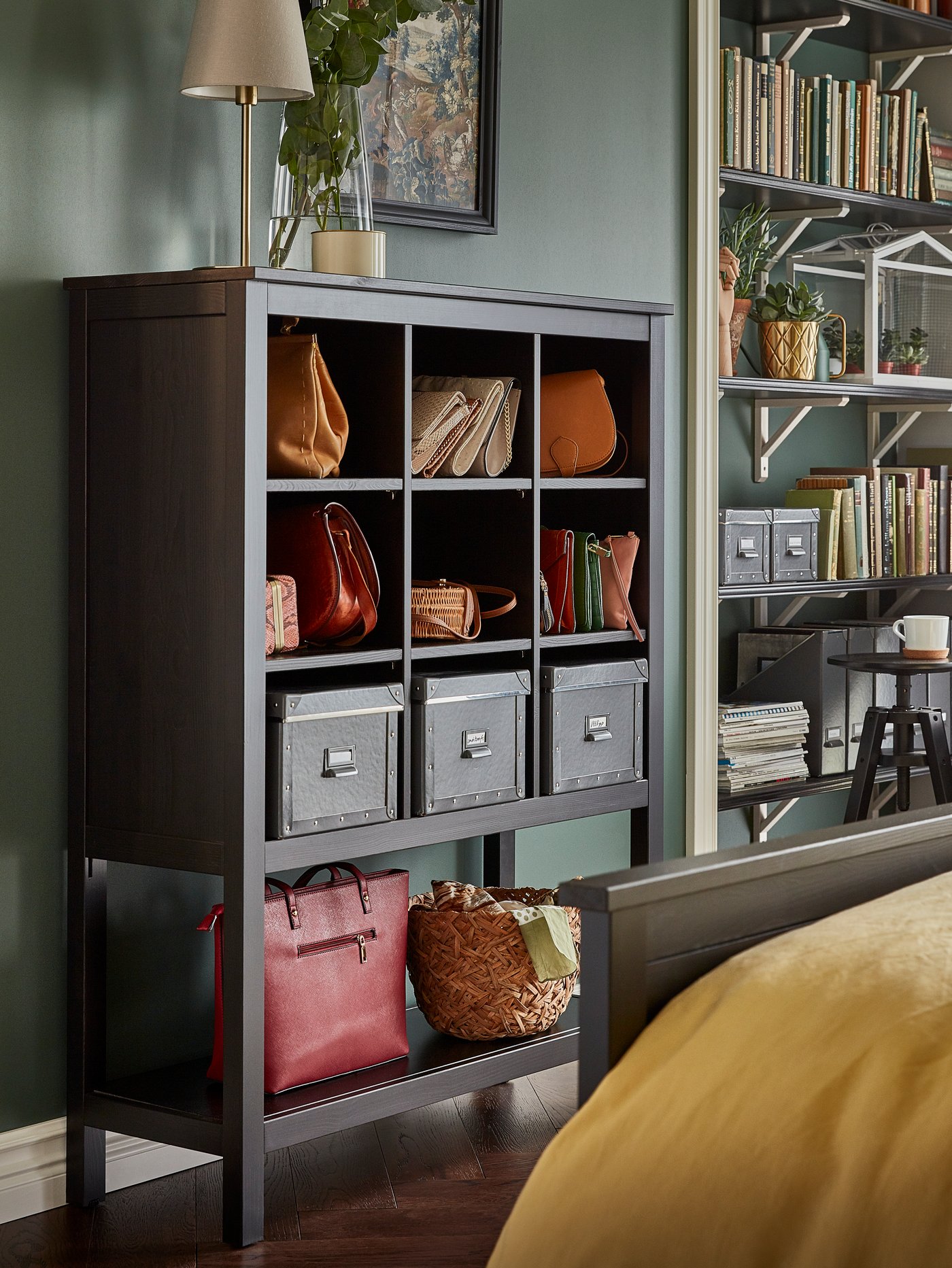 Bags and boxes sit on the shelves of a black-brown HEMNES storage unit which stands against the wall of a bedroom.