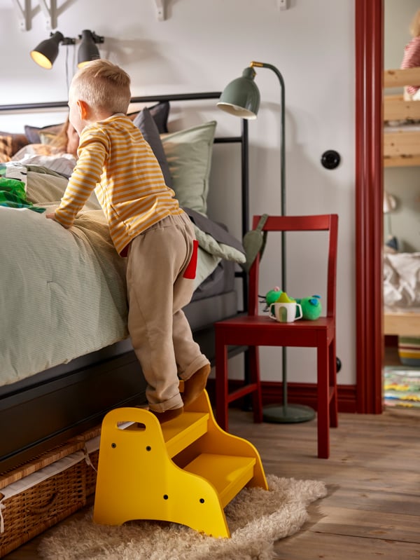 Small child climbing up to parents’ bed on yellow TROGEN children’s step stool. Red wooden chair used as bedside table.