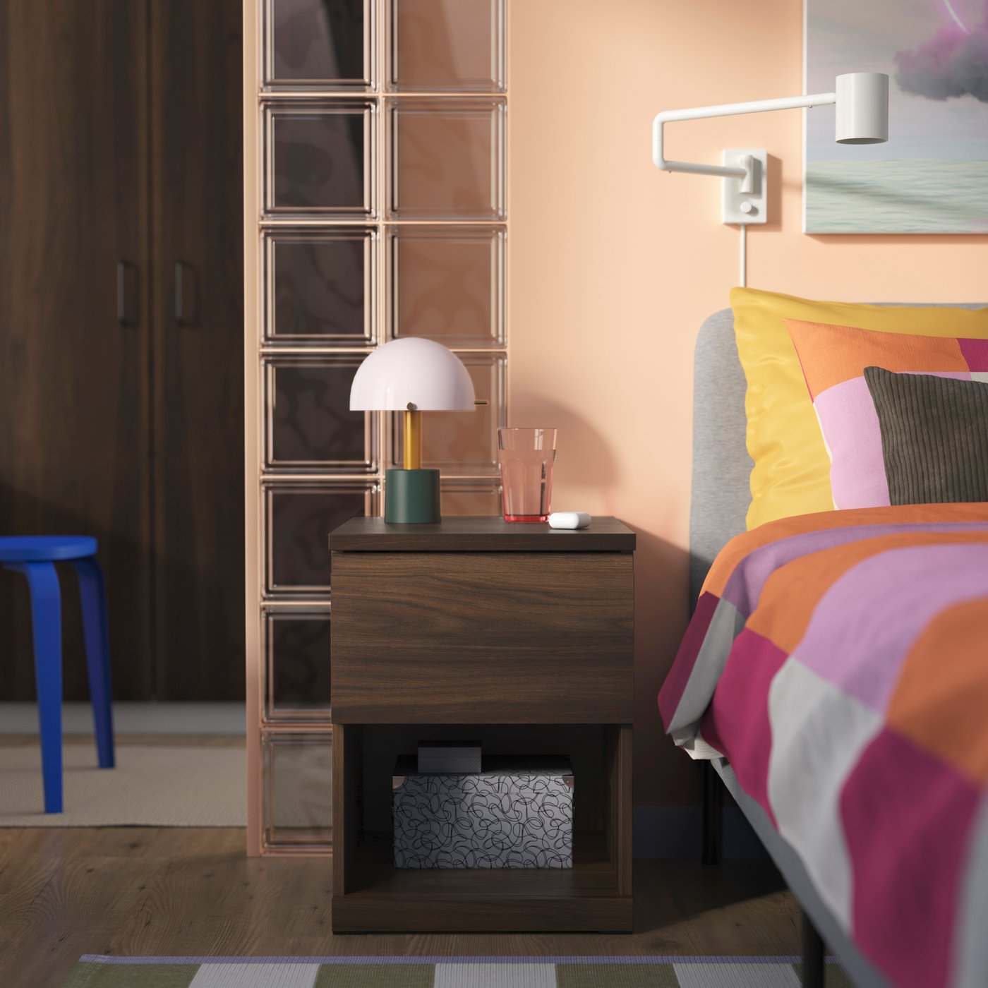 Ett sovrum i retrostil med ett mörkt trä-nattduksbord med en NÖDMAST lampa. Bredvid står en säng med färgglad randig bäddtextil i orange, rosa och lila toner. Väggarna är ljusa persikofärgade, och en vit vägglampa är monterad ovanför sängen.