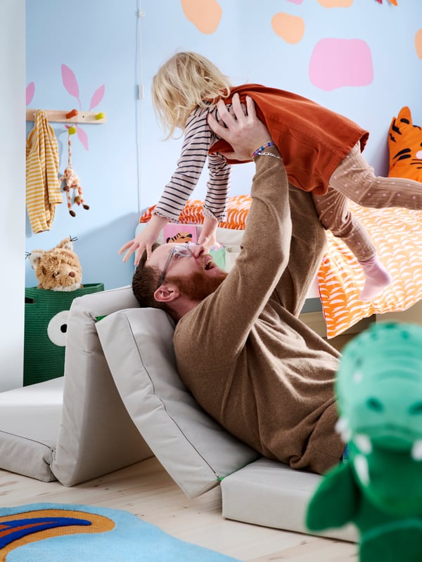 Father and child playing on SLÄKT pouffe/mattress on the floor, light blue walls behind and a hand puppet in the foreground.