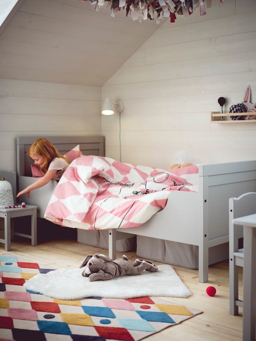 A girl lying in a grey SUNDVIK extendable bed covered with pink and white bed linen while reaching for a toy on her bedside table.