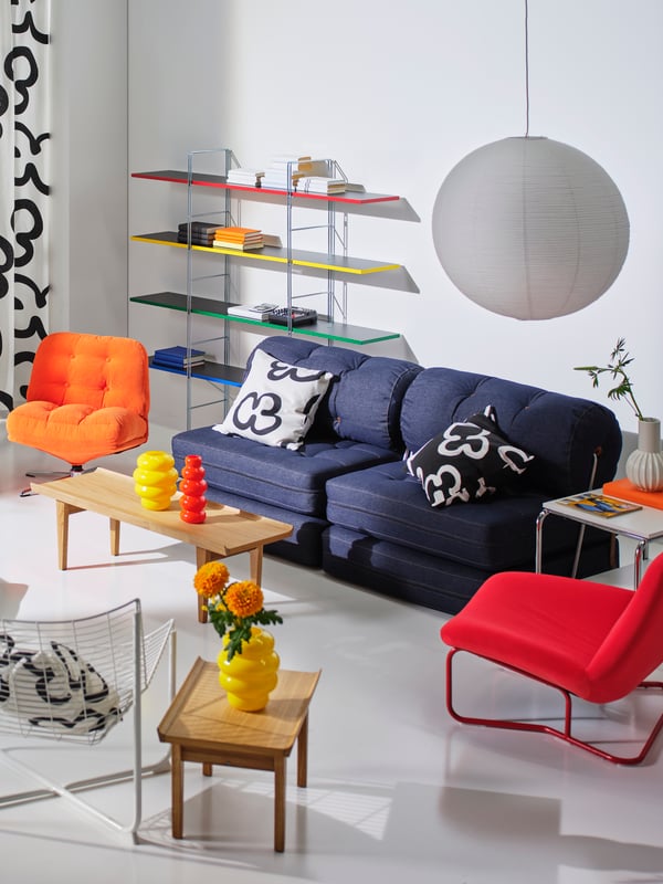 Modernes IKEA Wohnzimmer mit navyblauem Sofa und kontrastreichen schwarz-weißen Kissen. Farbliche Akzente setzen die orangefarbene und rote Sessel sowie die gelbe Vase auf dem Couchtisch. Die weiße Papierlampe und das bunte Wandregal unterstreichen den verspielten, kreativen Wohnstil.

