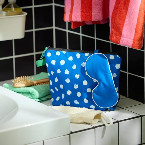 A blue SKOGSSALLAT toiletry bag with white dots and a blue eye mask on a white tiled shelf next to a bath tub. Green towels.