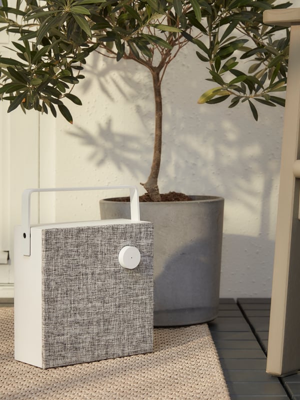 Enceinte Bluetooth carrée blanc/gris avec poignée disposée sur un tapis beige à côté d’une plante dans un pot gris.