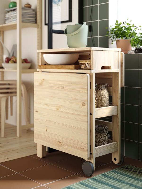 A wooden RESARÖ mobile drop-leaf table with storage is folded down and it has several kitchen items placed on the shelves.