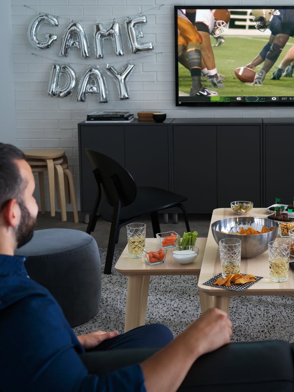 A man watches sport on TV in a living room, with drinks and snacks set out on LISABO tables.