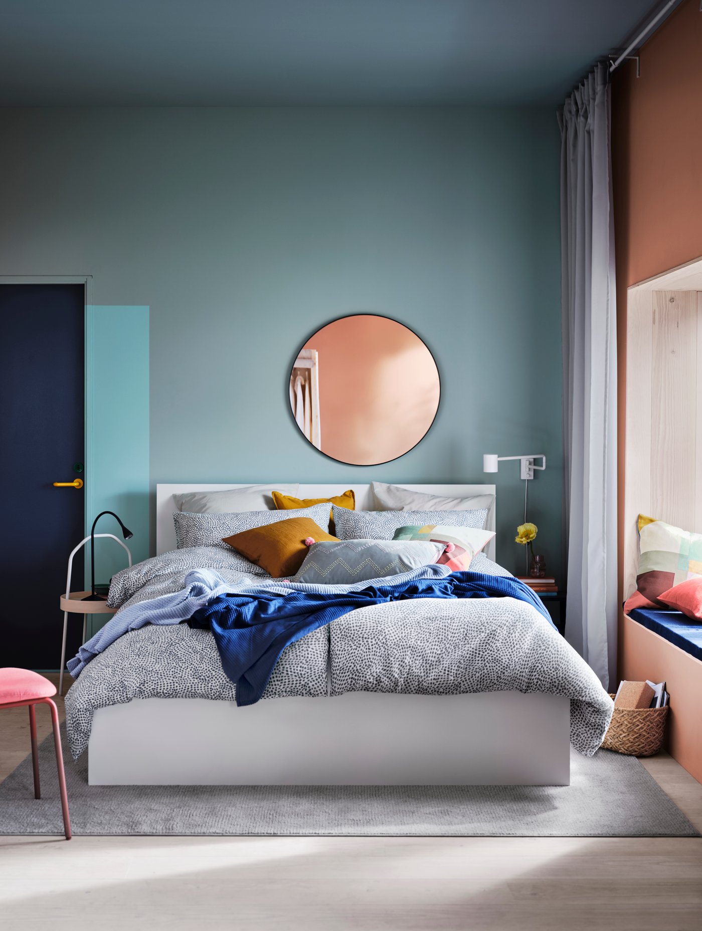 MALM white bedframe with spotted bedspreads in a cyan-blue room. A mirror hangs above the bed on the wall adjacent to the bed and a window sits right to the bed and a door to the left.
