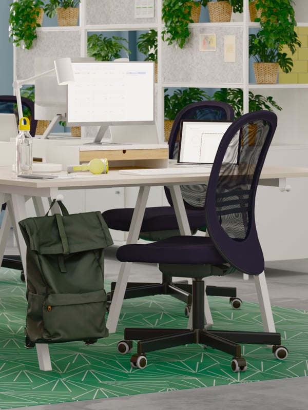 A white desk on a green rug and a white room divider with green plants behind it. Two black FLINTAN office chairs and a bag.