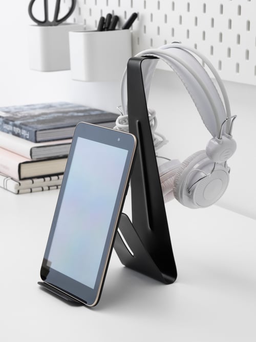 A tablet on a black MÖJLIGHET headset and tablet stand on a white desk with headphones hanging from it.