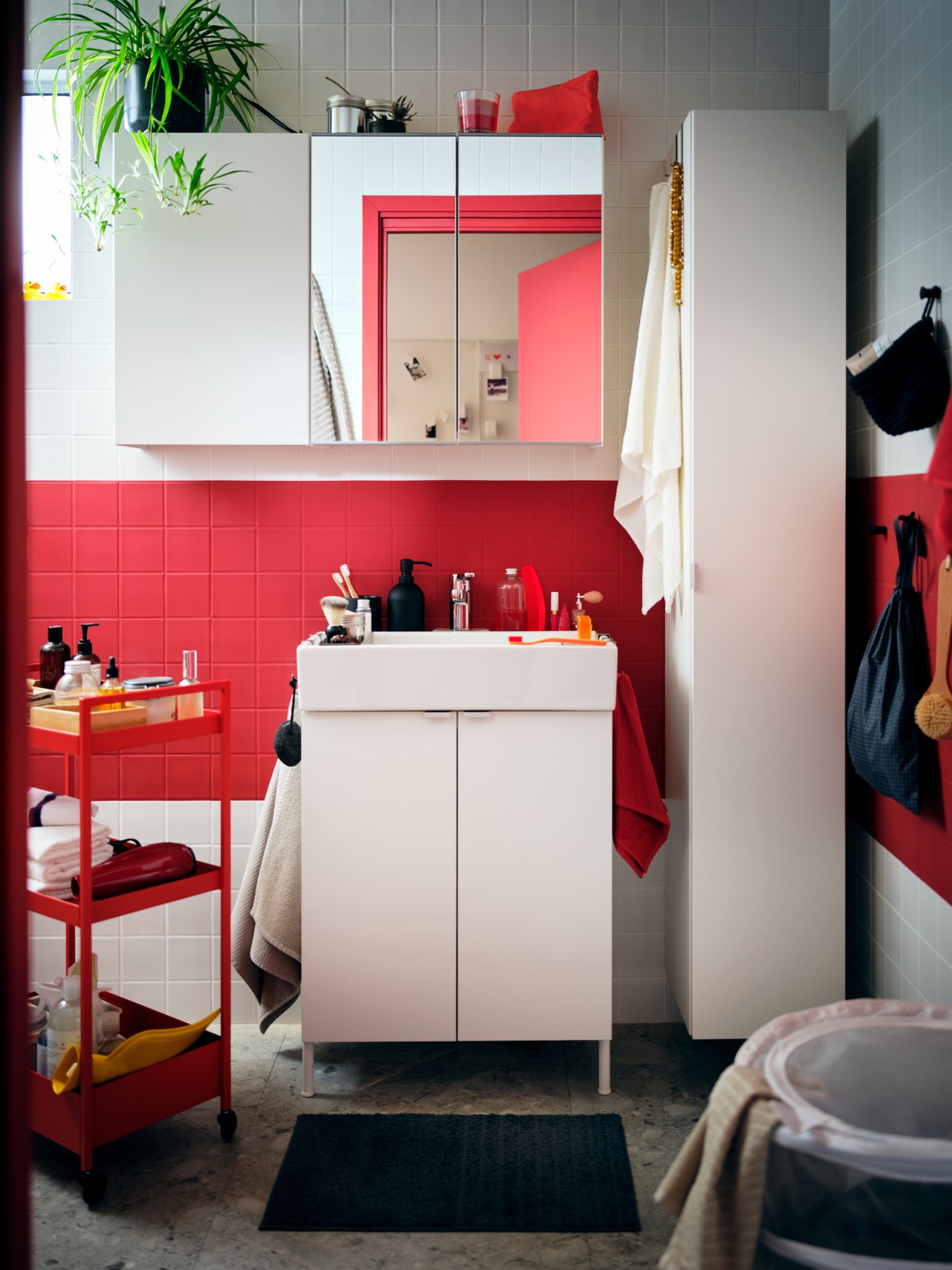 Un cuarto de baño con azulejos rojos y blancos, con gabinetes LILLÅNGEN blancos y un carrito NISSAFORS rojo con accesorios.