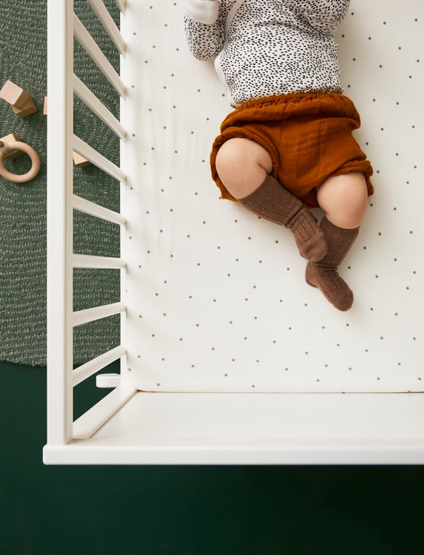 Bebis liggande i en spjälsäng med vita spjälor och en vit madrass med små prickar. Bebisen har på sig en grå långärmad tröja, bruna shorts och bruna knästrumpor. På den gröna mattan bredvid spjälsängen ligger några träleksaker.