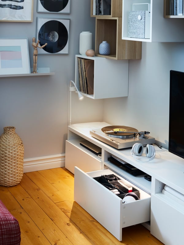 A white TV bench with an open and illuminated drawer, white wall cabinets, a small wall lamp and a decoration vase in bamboo.