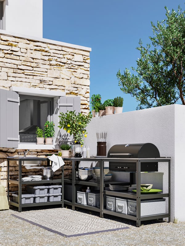A black outdoor kitchen by a window and wall, with a grill, sink, and shelves with tableware and cookware.