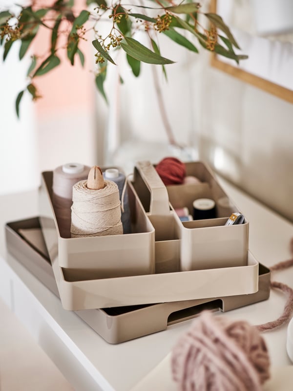 A grey-beige KUGGIS desk organiser set on a white surface holding different coloured sewing threads, with a plant overhead.