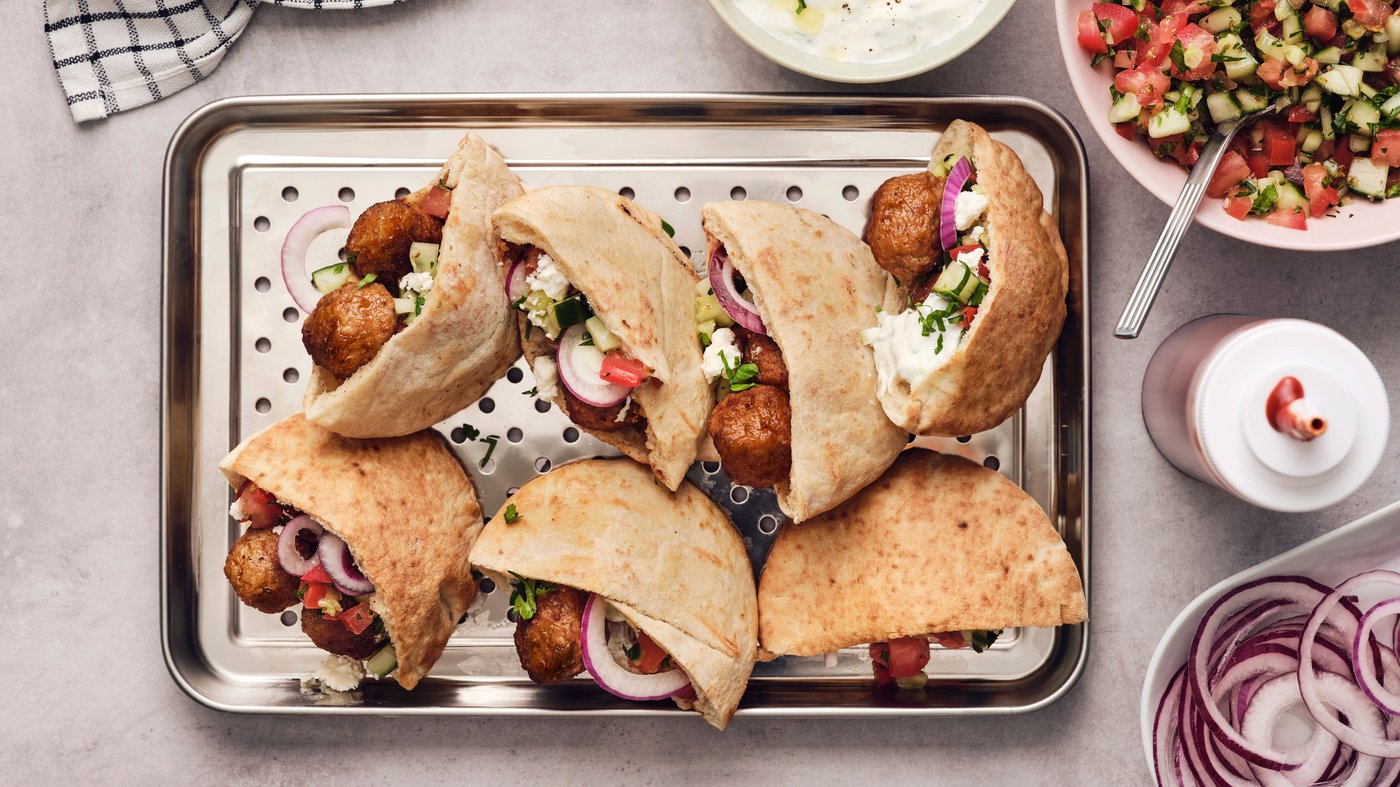 Pieces of pita bread, filled with meatballs, vegetables and tzatziki, are placed on a metal tray on a light grey worktop.