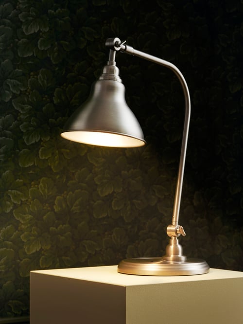 A lit work lamp with a round base support and two arms is placed on a cube. The background is a wall with leaf wallpaper.