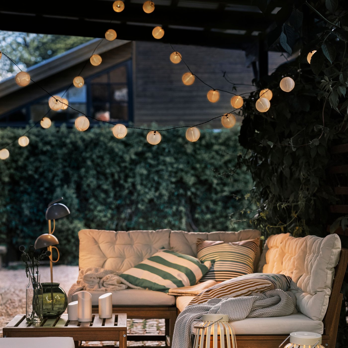 Mehrere Personen sitzen auf hellbraunen Sofas für draußen im Zwielicht auf einer Terrasse, die von einer LED-Lichterkette und zwei Standleuchten erhellt wird.