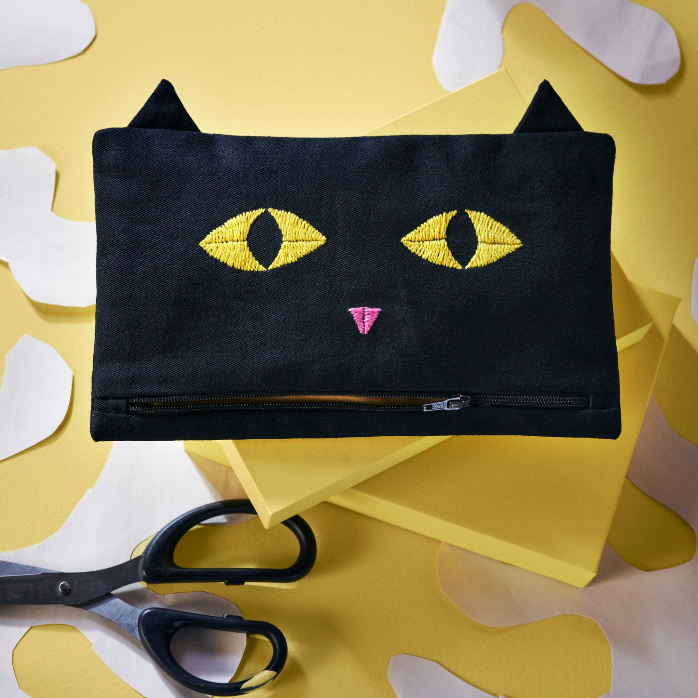 Černé pouzdro MÄVINN se vzorem kočky na žlutém stole a pod ním jsou nůžky. 