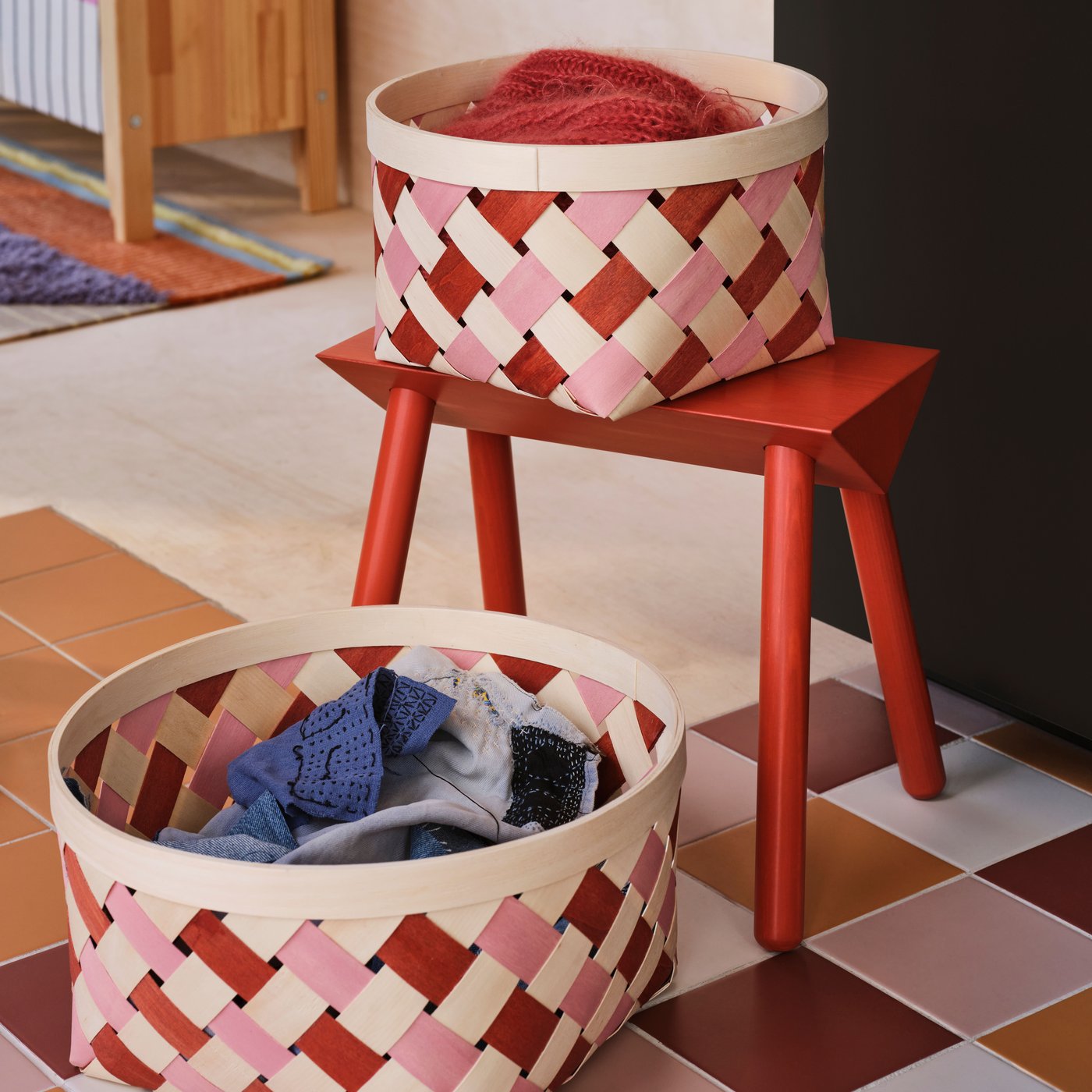 Two pink/orange OMMJÄNGE baskets containing textiles on a checkered floor next to a red stool.