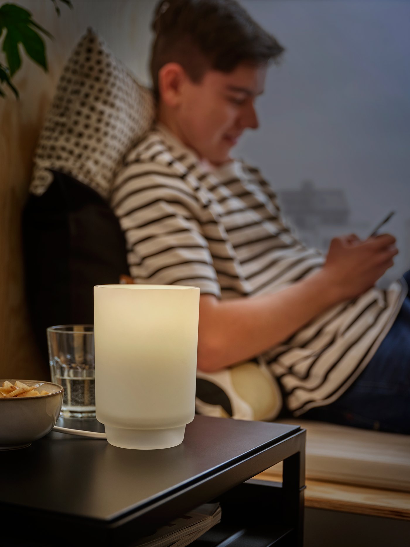 Lampu meja ISBRYTARE kaca berkabut memancarkan sinaran yang nyaman sambil seorang lelaki remaja berbaju kemeja berjalur menghantar mesej kepada rakan pada telefonnya.