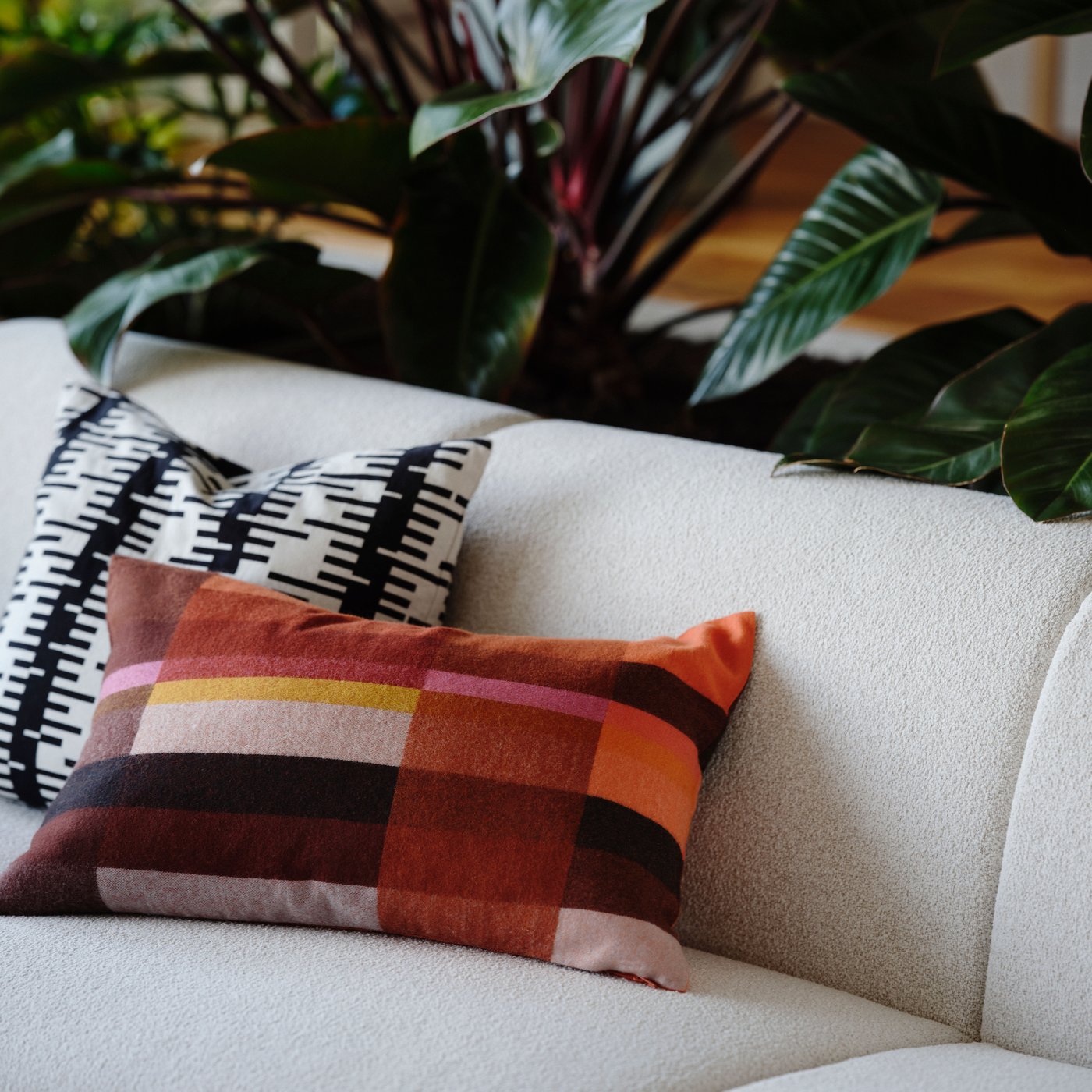 En soffa med två kuddar med olika STOCKHOLM 2025 kuddfodral står framför några växter.