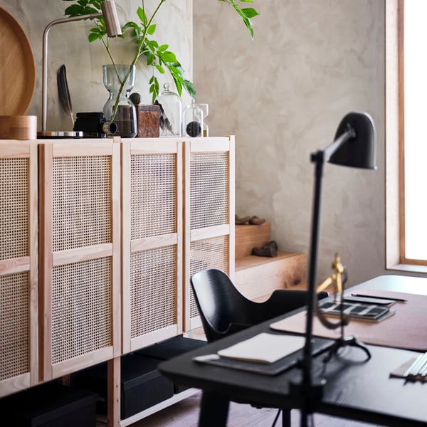 Ein modernes Arbeitszimmer mit minimalistischen Möbeln. Ein Rattanregal und eine schwarze Schreibtischlampe ergänzen den Raum, der von natürlichen Elementen wie Pflanzen und Holz akzentuiert wird. Der Tisch ist ordentlich mit Notizen und Schreibutensilien gedeckt, während ein großes Fenster für viel Tageslicht sorgt.