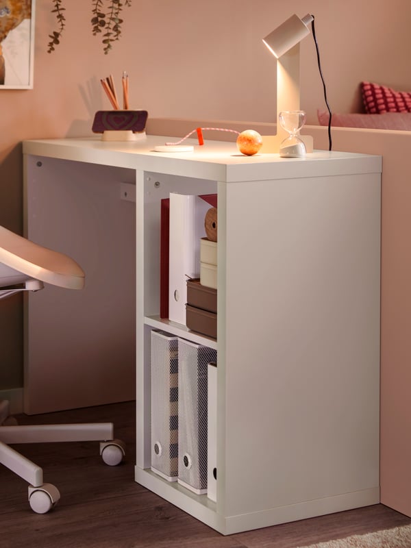 A white KALLAX desk with DRÖNJÖNS and KUGGIS magazine files, a lit, beige desk lamp and a white swivel chair.